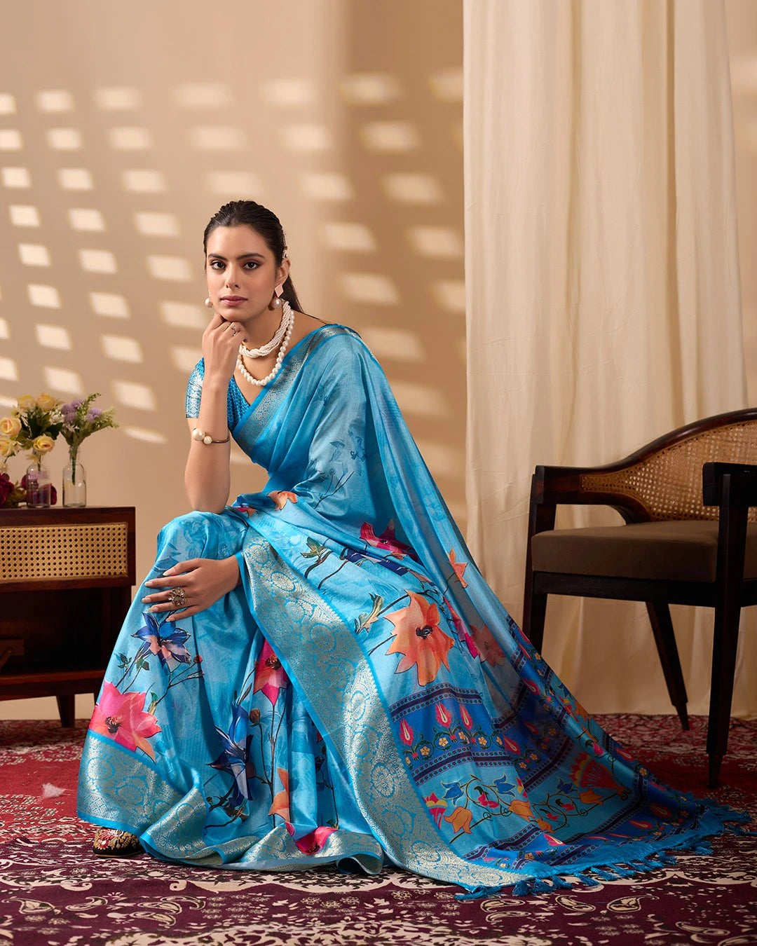 Woman in a blue saree with floral patterns sitting indoors.